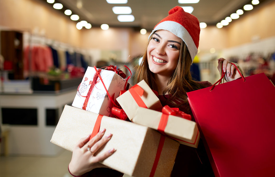 A person wearing a Santa hat smiles while holding several wrapped gifts and a red shopping bag in a store.