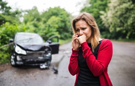 A concerned woman standing on a road near a damaged car after accident and making a phone call