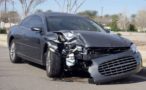 Silver car with severe front-end damage, including a crumpled hood and detached bumper, parked on a road near trees and grass.