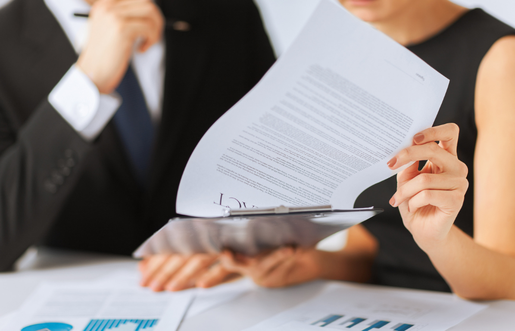 Two people in business attire review documents and graphs on a desk. One person holds a report on a clipboard, while the other appears to consider the information.