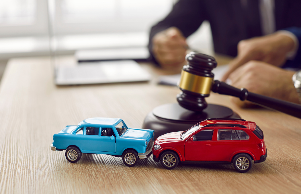 Two toy cars, blue and red, in a collision next to a gavel on a wooden desk, with a blurred figure in the background.