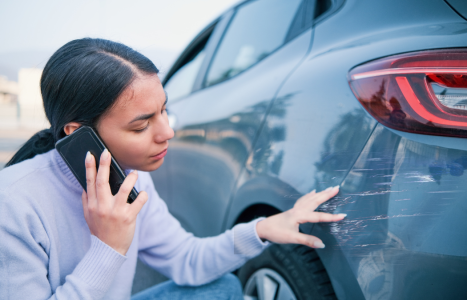 A woman on a phone call examines scratches on the side of a car.