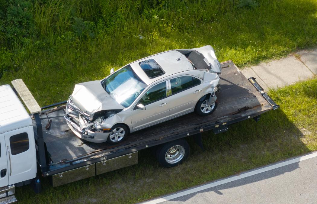 A damaged silver car with front and rear impact is secured on a flatbed tow truck on a roadside.