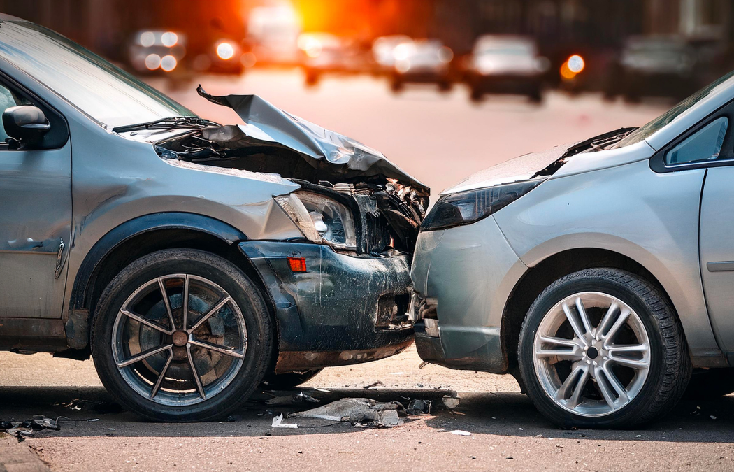 Two cars in a frontal collision on a city street. The front of both vehicles is damaged. Sunlight is visible in the background.