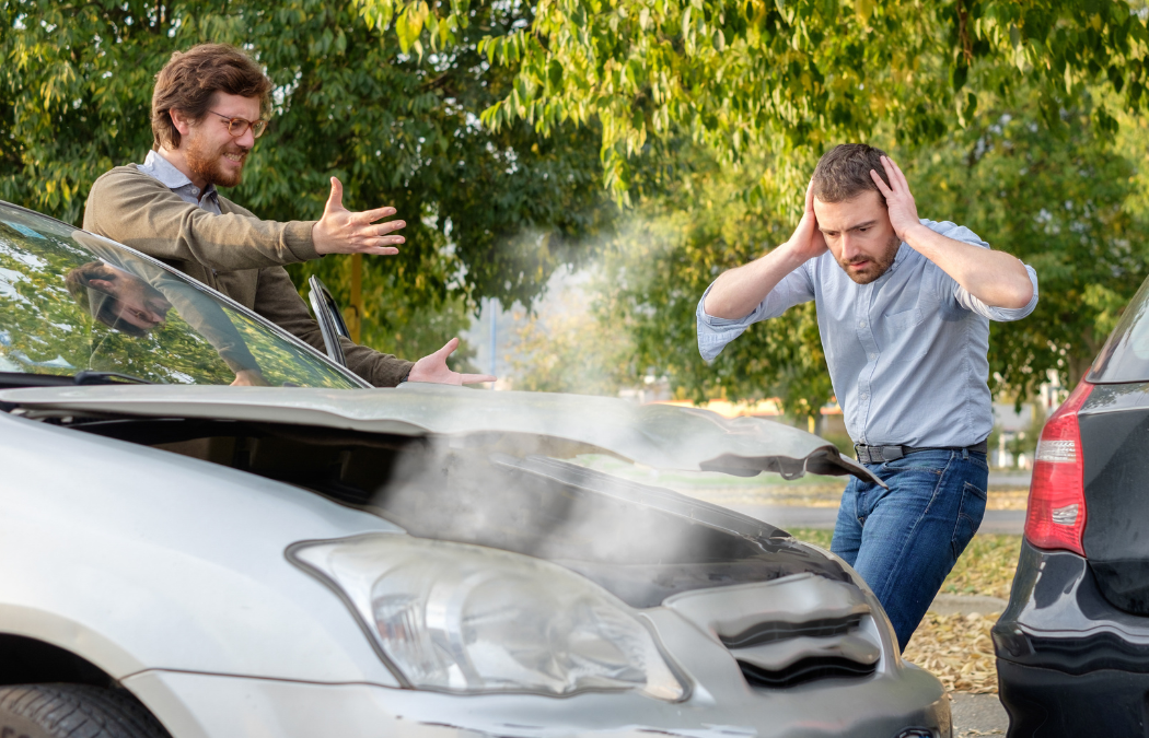 Two men stand next to a car with a damaged hood. One looks upset with hands on his head, while the other gestures with his hands. Steam is rising from the car. Trees are in the background.