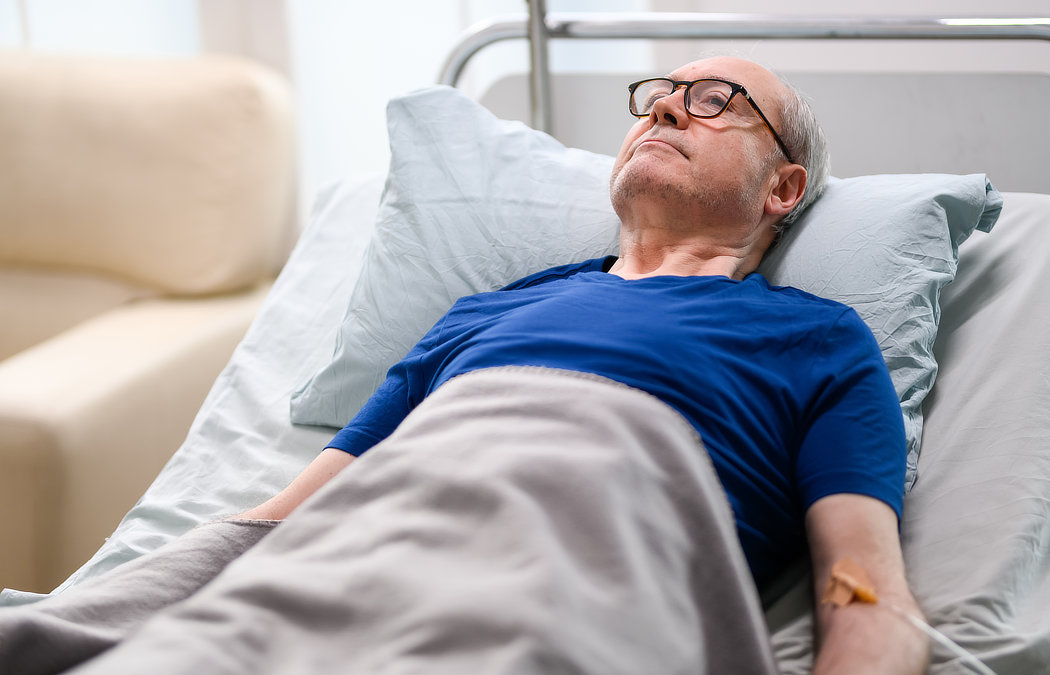 Elderly man with glasses lying in a hospital bed, wearing a blue shirt, covered with a gray blanket, and with an IV drip in his arm.