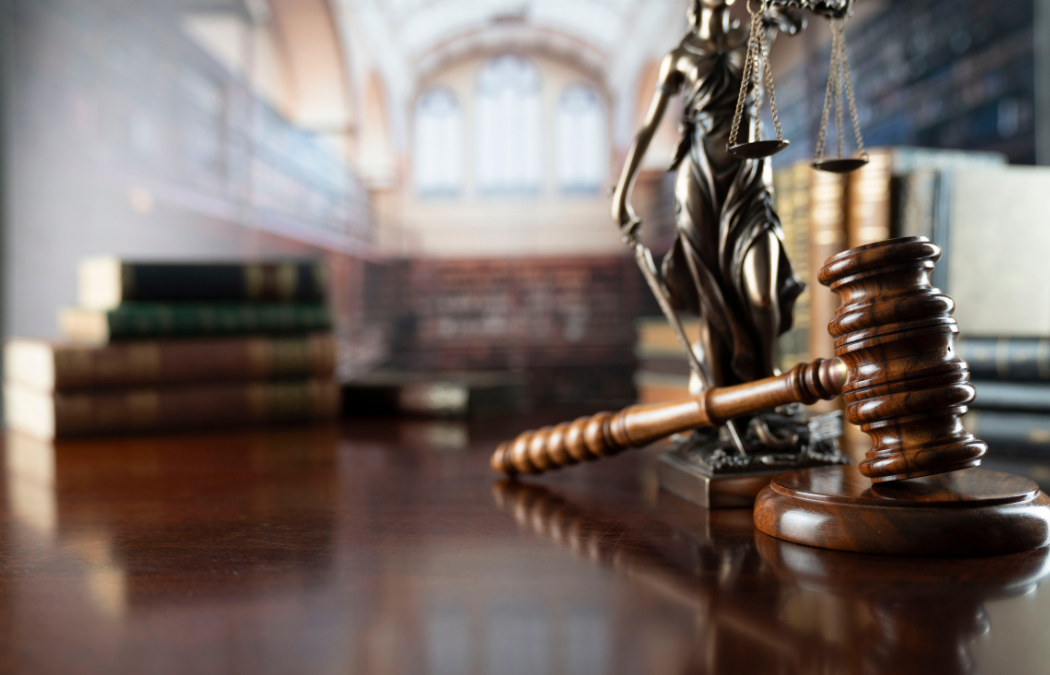 A gavel rests on a wooden desk near a statue of Lady Justice, with law books and blurred shelves in the background.