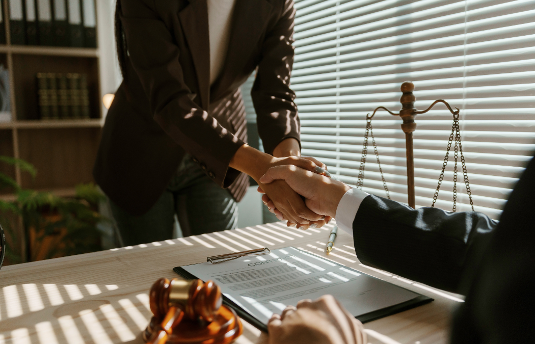 Two people shaking hands across a desk with a contract, scales of justice, and a gavel in an office setting.